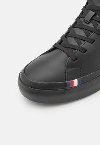 Zapatilla deportiva de cuero negro con textura suave, puntera redondeada y suela de goma con franjas azules, blancas y rojas en el lateral.