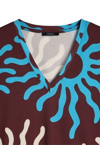 T-shirt z dekoltem w serek w brązowej kolorystyce. Posiada niebieskie i kremowe abstrakcyjne faliste wzory. Wykonany z gładkiego materiału o lekkiej teksturze.
