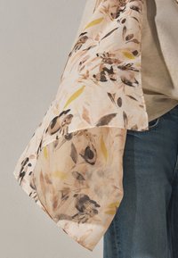Sciarpa leggera traslucida con motivo di conigli marroni e neri e foglie gialle, drappeggiata sulla spalla, indossata con top beige e jeans blu.
