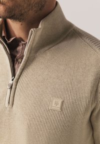 Beige gebreide trui met een hoge kraag en halve ritssluiting, met een subtiele geribbelde textuur en een vierkante logopatch op de borst.