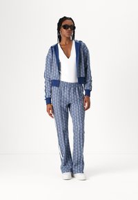 Tuta blu con motivo a fantasia, composta da una felpa con zip e pantaloni abbinati con strisce bianche ai lati. Il modello indossa occhiali da sole e calzature bianche.