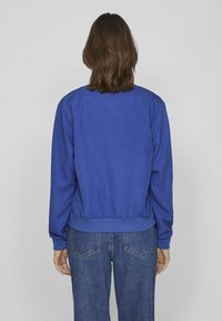 Chaqueta bomber azul con cintura y puños acanalados, textura suave y un diseño sencillo; vista desde atrás.