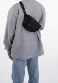 Svart midjeväska i slitstarkt tyg, med dragkedja och justerbar rem, bärs över en grå oversize-tröja och blåjeans.