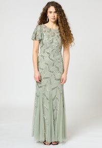 Langes olivengrünes Kleid mit paillettenbesetzten Blattmustern und kurzen, flatternden Ärmeln. Fließender, gestufter Tüllrock. Schlichte schwarze Sandalen.