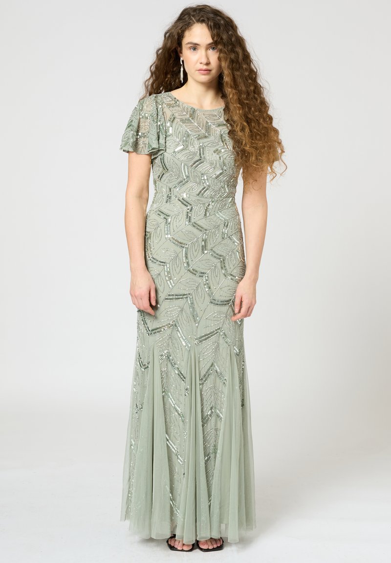 Langes olivengrünes Kleid mit paillettenbesetzten Blattmustern und kurzen, flatternden Ärmeln. Fließender, gestufter Tüllrock. Schlichte schwarze Sandalen.