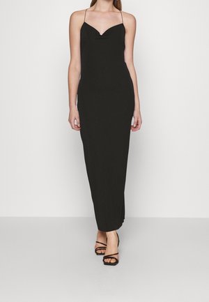 Frau trägt ein langes schwarzes Slip-Kleid mit dünnen Trägern und schwarze Riemchensandalen mit offenem Zehenbereich und steht vor einem schlichten hellen Hintergrund.