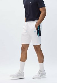 Muž v bílých sportovních šortkách s černými a modrými bočními pruhy, bílých teniskách, bílých ponožkách a tmavě modrém tričku s logem "BOSS".