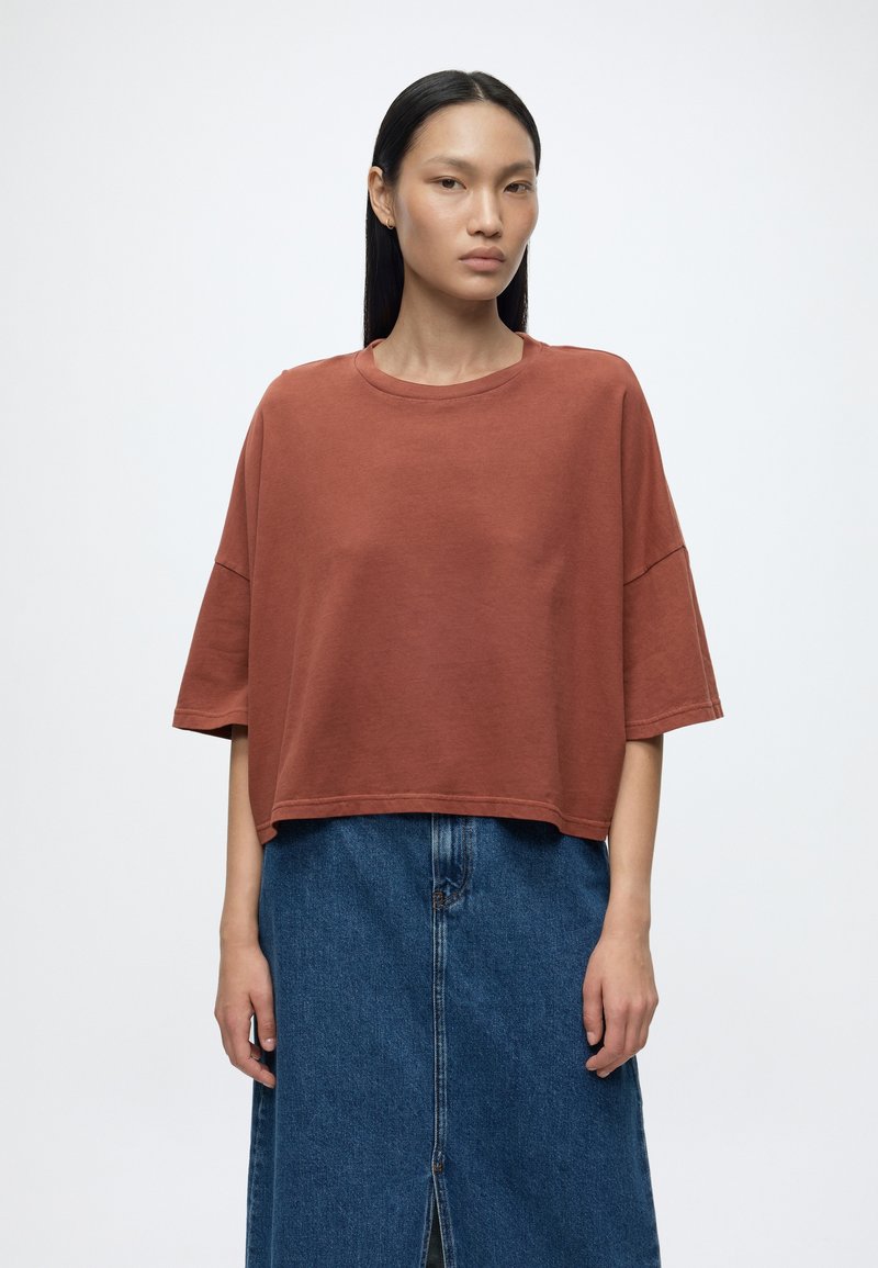 Femme aux cheveux noirs lisses portant un t-shirt ample oversize couleur rouille et une jupe en denim bleu taille haute avec une fente à l'avant.