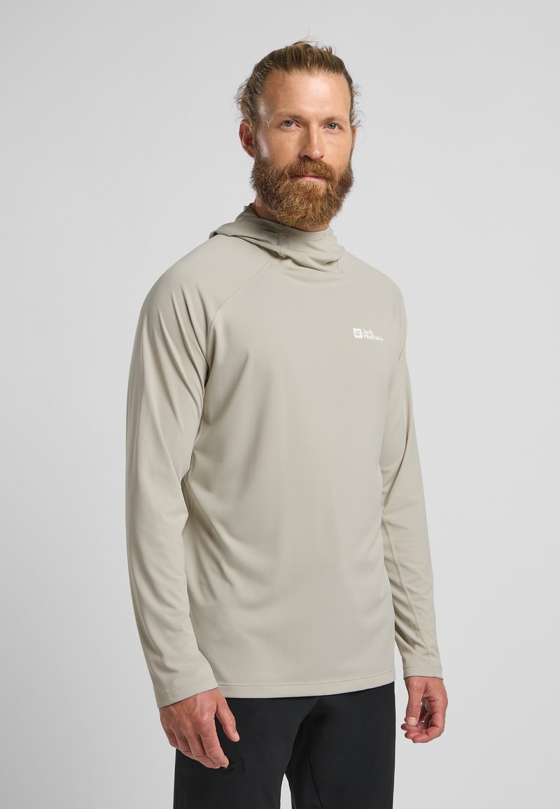 Chemise à manches longues beige avec capuche, fabriquée en tissu lisse, présentant un logo discret sur la poitrine gauche et une coupe décontractée.