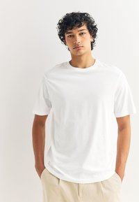 Valge puuvillast T-särk ümarate kaeluse ja lühikeste varrukatega. Sile pind, lahtine lõige, minimaalne brändimine ääres. Kandmisega beežid püksid.