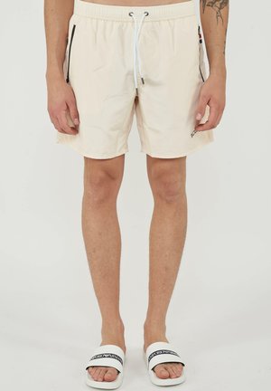 Short beige avec cordon de serrage et fermetures éclair latérales noires porté par une personne debout pieds nus dans des sandales slides blanches Emporio Armani.