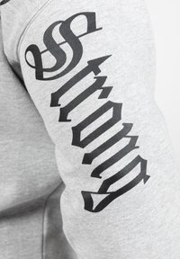 Grijze sweater met zwarte "Strong" tekst langs de mouw in een gestileerd lettertype, met een zachte, katoenachtige textuur.
