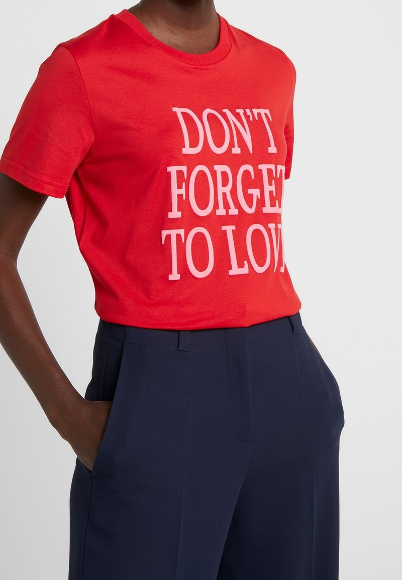Personne portant un t-shirt rouge avec un texte blanc « N'OUBLIEZ PAS D'AIMER » rentré dans un pantalon bleu marine, les mains dans les poches.