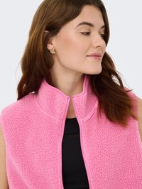 Roze fleecevest met een hoge kraag, een gestructureerd oppervlak en een ritssluiting aan de voorkant. Het model draagt gouden oorbellen, die een verfijnd accent toevoegen.