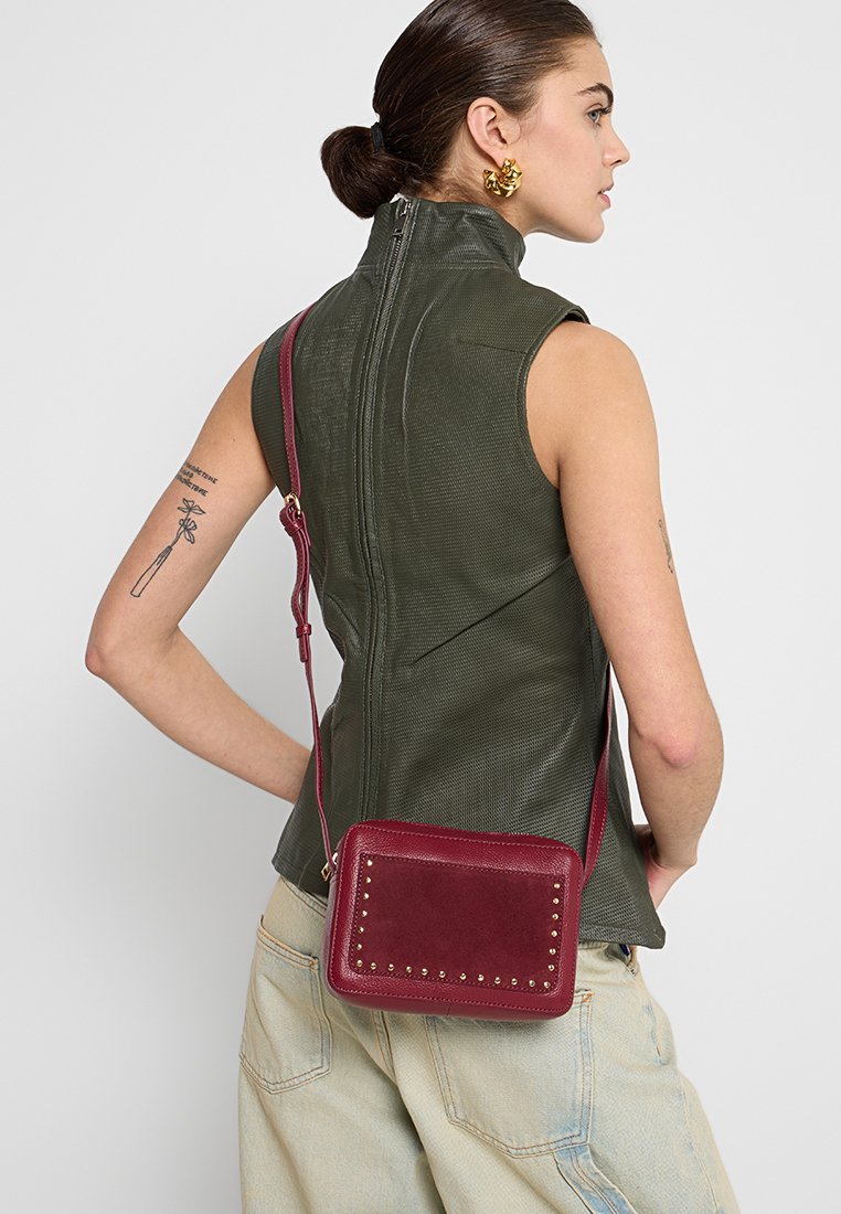 Burgundowa torba na ramię z miękkiej skóry, z ćwiekami, w prostokątnym kształcie i regulowanym paskiem. Nosi się ją na zielonym topie bez rękawów.