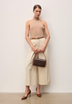 Naine, kes kannab beeži värvi kaelarätiga topi, laia säärega kreemikaid pükse, pruuni vööga, pruunide kõrgete kontsadega, hoides pruuni mustriga käekotti ühtlase tausta ees.