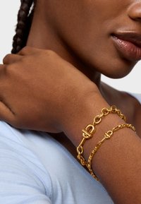 Bransoletki z metalowego złota noszone na nadgarstku, z charakterystycznymi przeplatającymi się ogniwami i zapięciem. Odcień skóry to głęboki brąz, na tle jasnoniebieskiej bluzki.