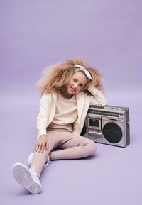 Dijete s kovrčavom plavom kosom, odjeveno u svijetlosivi pulover i ružičaste tajice, sjedi pored srebrnog boomboxa na svijetlo ljubičastoj pozadini.