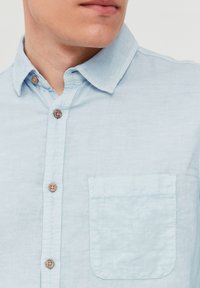 Camisa de lino en azul claro con cuello abotonado, botones de madera y un solo bolsillo en el pecho con una solapa redondeada. Textura suave.