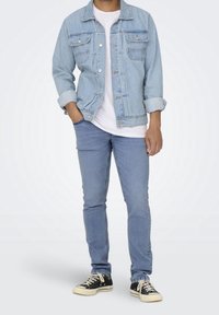 Chaqueta de mezclilla azul claro con frente abotonado, mangas largas, bolsillos en el pecho, bordes desgastados, combinada con jeans claros a juego y zapatillas negras.