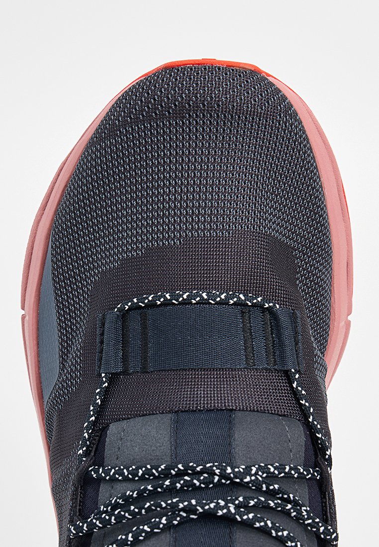Donkerblauwe sneaker met een gestructureerde gebreide bovenkant, zwart-witte veters, brede band over de middenvoet en roze rubberen zolen.