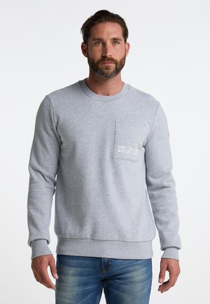 Lys grå sweatshirt i blødt materiale, med rund hals, lange ærmer og en brystlomme med hvid tekst. Parret med jeans.