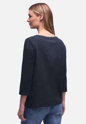 Kvinde med glat blondt hår iført navyblå top med trekvartærmer og blå jeans, set bagfra mod en hvid baggrund.