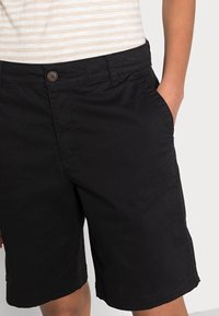 Osoba v černých kraťasech po kolena a béžovo-bílém pruhovaném tričku, s jednou rukou v kapse.