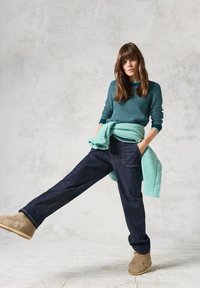 Jean larges bleu foncé associés à un pull texturé bleu sarcelle, porté autour de la taille. Des bottines en daim beige complètent le look décontracté.