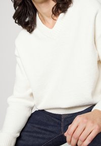 Bílý svetr s výstřihem do V z měkké, texturované látky, s žebrovanými manžetami a lemem, doplněný tmavými džínami.