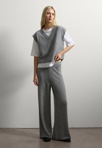 Donna bionda con pantaloni grigi a maglia larghi e un maglione senza maniche a scollo a V sopra una maglietta bianca, in piedi contro un muro bianco semplice.