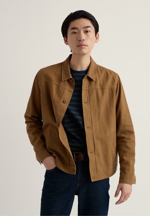 Joven con una chaqueta marrón abotonada sobre una camisa a rayas verde oscuro y azul marino, con vaqueros azul oscuro, de pie frente a un fondo liso.