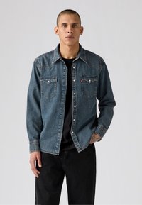Cămașă din denim de culoare bleumarin închis, cu deschidere cu nasturi, guler ascuțit, două buzunare pe piept și cusături contrastante. Purtată peste un top negru.
