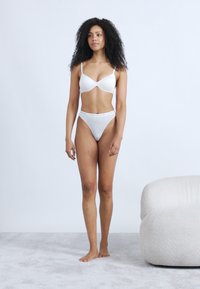 Λευκό επενδεδυμένο σουτιέν και γκρι ψηλόμεσο στρινγκ, απαλό ύφασμα, μίνιμαλ σχεδιασμός, το μοντέλο στέκεται σε ανοιχτόχρωμο χαλί, ουδέτερο φόντο.