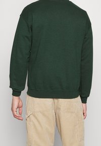 Zelená mikina s kulatým výstřihem a dlouhými rukávy, s žebrovanými manžetami a spodním lemem. Nositá přes béžové kordové kalhoty s bočními kapsami.