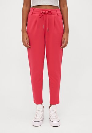 Vrouw in hooglopende, roze taps toelopende broek met een trekkoord in de taille en witte hoge sneakers, staande tegen een effen achtergrond.