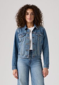 Giacca in denim blu con chiusura a bottoni, tasche laterali e leggeri dettagli consumati. Presenta un colletto classico e una linea sagomata. Abbinata a jeans a vita alta.