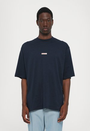 Marinblå oversized t-shirt med korta ärmar, rund halsringning och en liten röd och vit logotyp på bröstet. Mjuk bomullstextur.