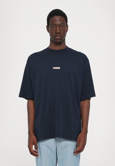 Camiseta de gran tamaño en azul marino con mangas cortas, escote redondo y un pequeño parche de logo en rojo y blanco en el pecho. Textura de algodón suave.