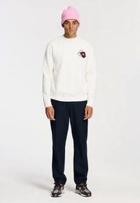 Kreminės spalvos džemperis su gėlių grafika, derinamas su tamsiai mėlynomis kelnėmis ir įvairiaspalviais sportbačiais. Šviesiai rožinė mezginė kepurė papildo aprangą.