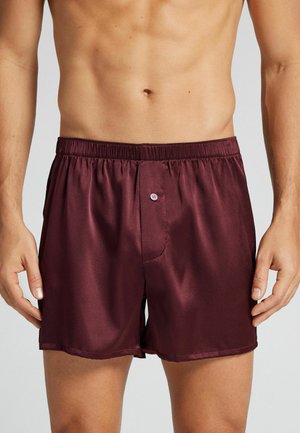 Un homme portant un boxer en satin rouge foncé brillant avec une ceinture élastique et un bouton à l'avant.