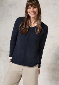Marineblauwe wollen trui met een V-hals, voorzien van ribgebreide manchetten en zoom. Gedragen over een wit overhemd met kraag, aangevuld met beige broeken.