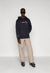 Marineblauwe hoodie met roze logo, capuchon met trekkoord, geribbelde manchetten, gecombineerd met beige broek en zwarte schoenen. Effen achtergrond.