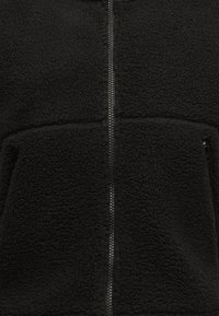 Svart fleecejakke med glidelås foran, teksturert overflate, to sidelommer og et enkelt design.