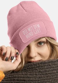 Pink ribbed beanie with a rectangular patch displaying white text "DIE MIT DEM HUND GEHT." Soft texture, fitted design, worn slightly above the brow.