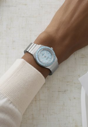 Handgelenk mit einer Timex-Uhr mit silbernem Metallarmband, die ein hellblaues Zifferblatt und weiße Stundenmarkierungen aufweist, liegend auf einer strukturierten beigefarbenen Oberfläche.