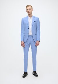 Fato azul claro com um corte ajustado, apresentando um fecho de um botão, lapelas com recortes e dois bolsos frontais. Usado com uma camisa branca e sapatos pretos.