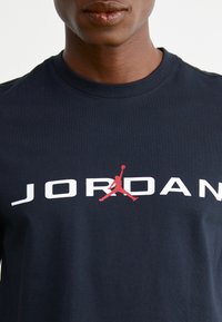 Jūras zils kokvilnas T-krekls ar lielu baltu tekstu "JORDAN" un sarkano siluetu logotipu centrā, apaļa dekoltē un īsām piedurknēm.