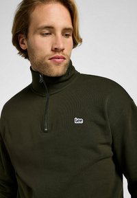 Pullover verde scuro con mezza zip in un tessuto morbido, con un piccolo logo bianco "Lee" sul petto. Presenta un colletto alto e una texture liscia.