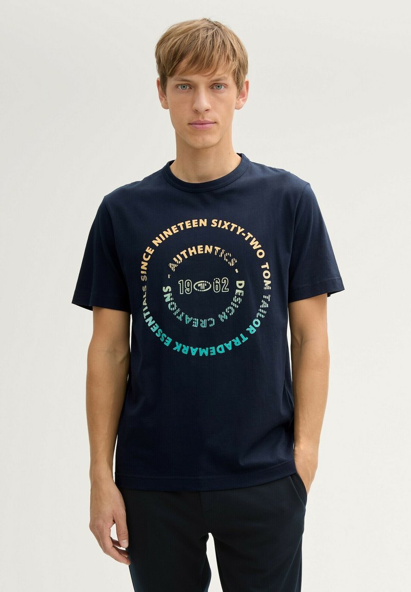 T-shirt en coton bleu marine avec un design graphique circulaire contenant du texte de différentes couleurs : or, turquoise et blanc. Manches courtes, col rond.